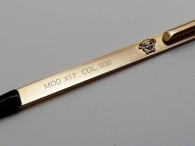 Puede incluir: Primer plano de una varilla de montura de gafas dorada. La varilla presenta el texto "MOD. X17 COL. 030" y un emblema de cabeza de Medusa. La varilla tiene una punta negra y una bisagra dorada.