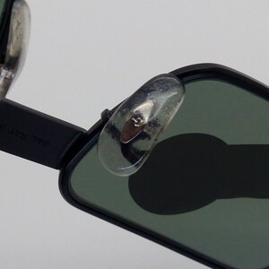 May include: Close-up of black sunglasses with green lenses. The sunglasses have a rectangular shape and silver nose pads. The brand name "B&L RAY-BAN" is visible on the arm of the sunglasses.