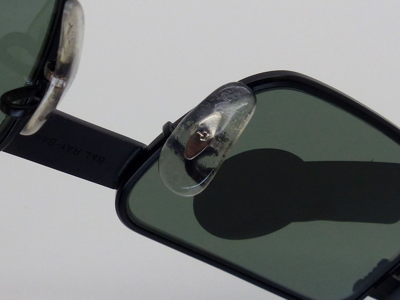 May include: Close-up of black sunglasses with green lenses. The sunglasses have a rectangular shape and silver nose pads. The brand name "B&L RAY-BAN" is visible on the arm of the sunglasses.