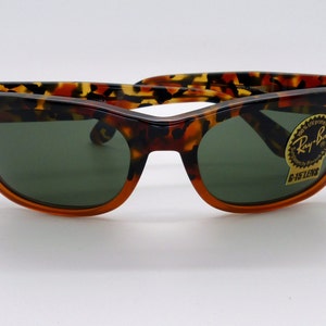Peut inclure: Lunettes de soleil à motif écaille de tortue avec verres verts. Les montures sont marron foncé et orange. Les lunettes de soleil ont un autocollant Ray-Ban sur le verre.