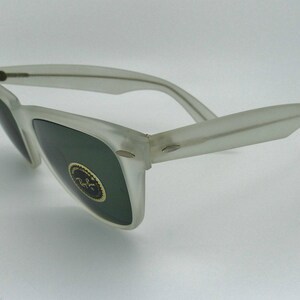 May include: Frosted white sunglasses with dark green lenses. The sunglasses have a classic design with a rectangular frame and curved temples. A gold emblem is visible on the lens. The sunglasses are angled, showing the side view.
