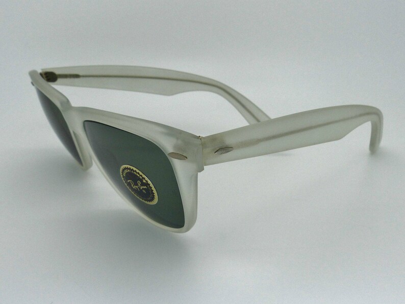 May include: Frosted white sunglasses with dark green lenses. The sunglasses have a classic design with a rectangular frame and curved temples. A gold emblem is visible on the lens. The sunglasses are angled, showing the side view.