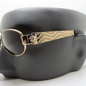 Puede incluir: Gafas de sol doradas con lentes oscuras. Las patillas presentan un diseño ondulado y un acento rectangular con un emblema de cabeza de Medusa. Las gafas de sol se exhiben en una cabeza de maniquí gris.
