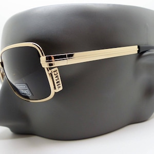 Puede incluir: Gafas de sol rectangulares con montura dorada y lentes oscuras, expuestas sobre una cabeza de maniquí gris oscuro. Las gafas de sol llevan el nombre de la marca "Versace" en la montura. El diseño es elegante y moderno.