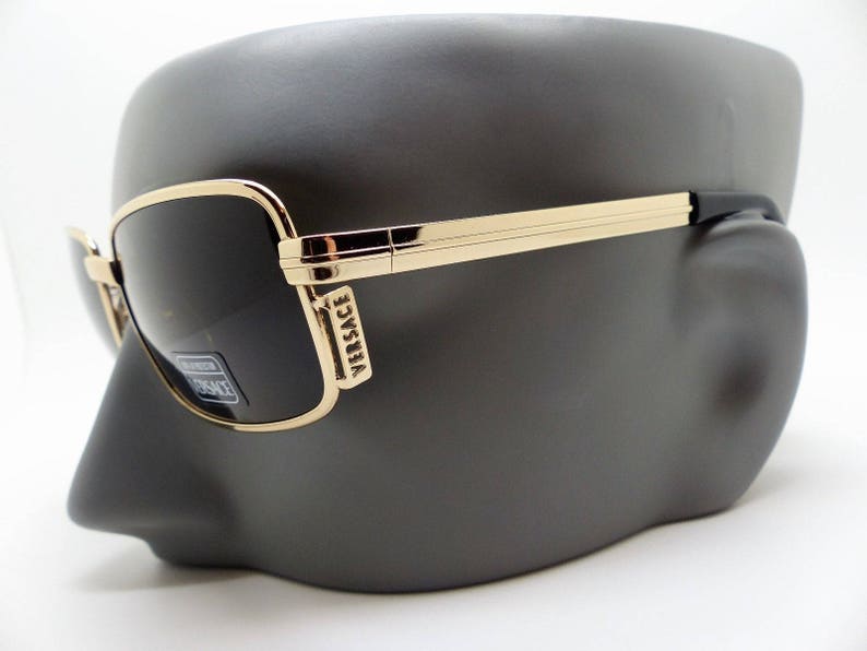 Puede incluir: Gafas de sol rectangulares con montura dorada y lentes oscuras, expuestas sobre una cabeza de maniqu&iacute; gris oscuro. Las gafas de sol llevan el nombre de la marca "Versace" en la montura. El dise&ntilde;o es elegante y moderno.