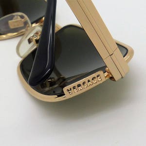 Puede incluir: Primer plano de gafas de sol con montura dorada y lentes oscuras. Las gafas de sol tienen una montura de metal dorado y patillas negras. El nombre de la marca "VERSACE" es visible en la montura.