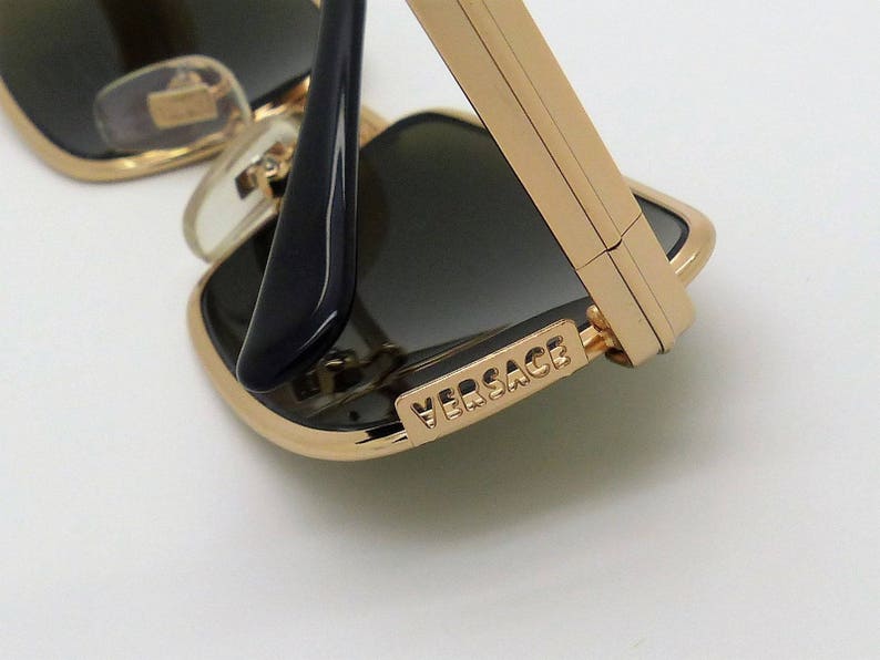 Puede incluir: Primer plano de gafas de sol con montura dorada y lentes oscuras. Las gafas de sol tienen una montura de metal dorado y patillas negras. El nombre de la marca "VERSACE" es visible en la montura.