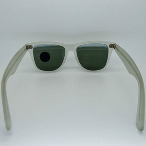 May include: Off-white sunglasses with dark green lenses. The sunglasses have a classic rectangular frame and temples that extend outward. The lenses are slightly reflective, and the frame is a matte finish.