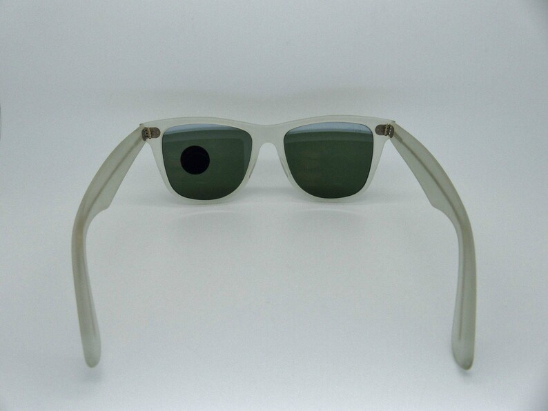 May include: Off-white sunglasses with dark green lenses. The sunglasses have a classic rectangular frame and temples that extend outward. The lenses are slightly reflective, and the frame is a matte finish.