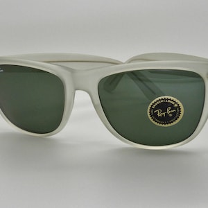May include: Frosted white sunglasses with dark green lenses. The Ray-Ban logo is visible on the left lens and on a circular sticker on the right lens. The sunglasses have a classic, retro design.