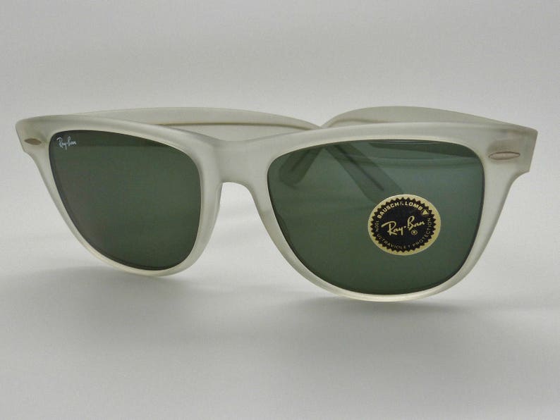 May include: Frosted white sunglasses with dark green lenses. The Ray-Ban logo is visible on the left lens and on a circular sticker on the right lens. The sunglasses have a classic, retro design.