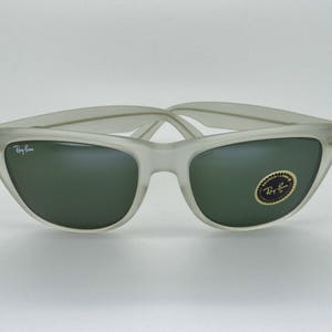 May include: Frosted gray sunglasses with dark green lenses. The Ray-Ban logo is visible on the left lens and the frame. A gold and black sticker is on the right lens. The sunglasses have a classic, retro design.