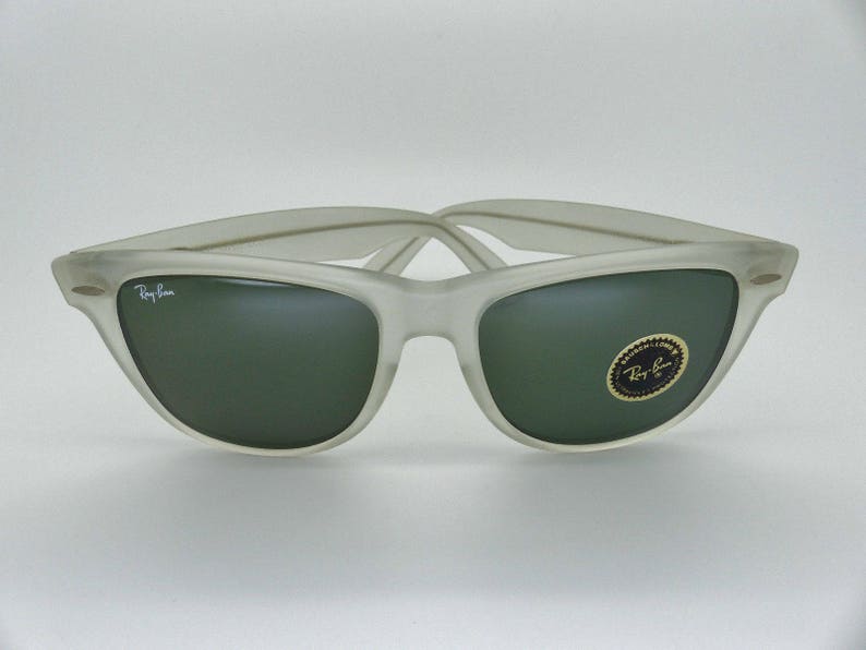 May include: Frosted gray sunglasses with dark green lenses. The Ray-Ban logo is visible on the left lens and the frame. A gold and black sticker is on the right lens. The sunglasses have a classic, retro design.