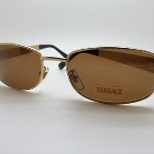Puede incluir: Gafas de sol doradas con lentes marrones. Las monturas rectangulares presentan un puente y varillas doradas, con bisagras negras. La marca "VERSACE" es visible en una etiqueta en la lente.
