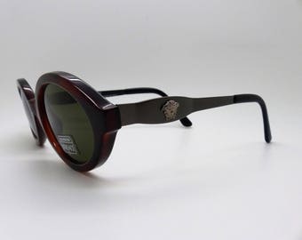 May include: Brown oval sunglasses with dark green lenses. The frames transition from a dark brown to a lighter brown. The temples are dark gray with a silver emblem. The arms are black.