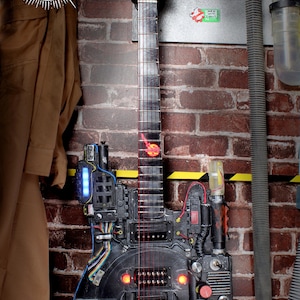 Könnte beinhalten: Eine maßgefertigte schwarze E-Gitarre mit roten Saiten und orangefarbenen Lichtern. Die Gitarre ist stark mit verschiedenen elektronischen Komponenten und Drähten modifiziert, die einem Requisit aus einem Science-Fiction-Film ähneln. Die Kopfplatte hat ein schwarz-weißes Design. Der Hintergrund ist eine Backsteinmauer.