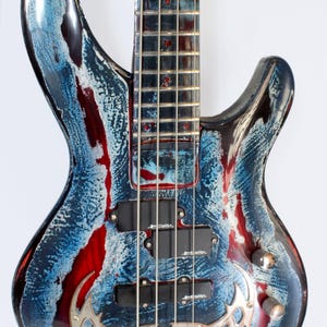 Könnte beinhalten: Eine individuelle E-Bassgitarre mit einem einzigartigen Finish. Der Korpus weist ein abstraktes Design in Blau, Rot und Weiß auf. Der Bass hat vier Saiten, schwarze Tonabnehmer und eine silberne Brücke. Kopfplatte und Korpus haben ein glänzendes Finish.