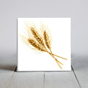 May include: A square ceramic tile featuring a watercolor illustration of wheat stalks in shades of gold and brown. The wheat is arranged in a small bundle against a white background.