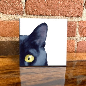 May include: Square art print featuring a watercolor illustration of a black cat's eye and ear. The cat's eye is yellow, and the fur is dark blue. The artwork is displayed on a wooden surface with a brick background.
