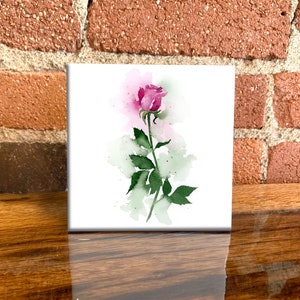 May include: Square white canvas art featuring a watercolor painting of a pink rose with green leaves and stem. The background has soft green and pink watercolor washes. The artwork is displayed on a wooden surface with a brick wall backdrop.