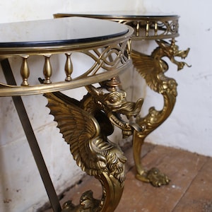 May include: Two antique brass side tables. Each table has a round top with a black border and ornate brass dragon legs. The dragon legs have detailed wings and a curved design. The tables are set against a white wall.