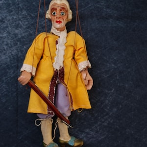 May include: A vintage marionette doll in a yellow coat with white trim, purple trousers, and blue and gold shoes. The doll has a painted face with red lips and holds a wooden rod. The strings are visible, indicating it is a puppet, a traditional toy.