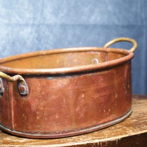 May include: An oval-shaped copper basin featuring a smooth, weathered metallic surface with visible patina and oxidation. The vessel includes two curved brass handles securely attached to the sides with rivets, providing a functional grip.
