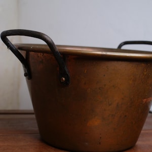 May include: A copper bucket with two black handles. The bucket has a rounded shape and a slightly aged appearance. The handles are attached to the sides with rivets. The bucket sits on a wooden surface.