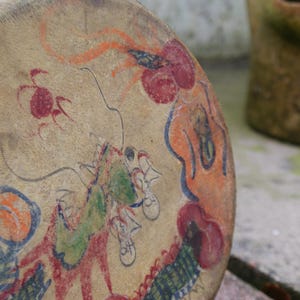 May include: A round, tan-coloured drum with a painted design of a green dragon, red flowers, and orange and red swirls. The drum is made of leather and has a wooden frame.