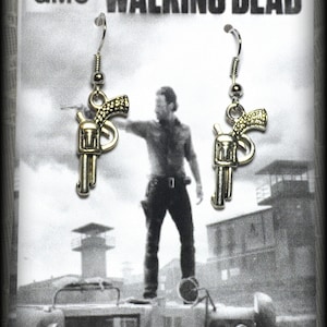 May include: A pair of silver earrings featuring a gun design. The earrings are on a white card with the text "The Walking Dead" and "AMC" printed on it.