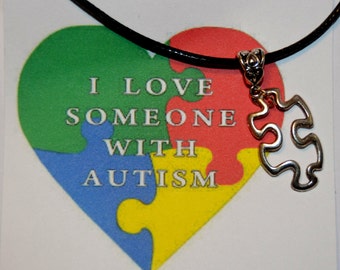 May include: A black cord necklace with a silver puzzle piece pendant. The necklace is displayed on a white background with a colorful heart-shaped design that reads "I love someone with autism".
