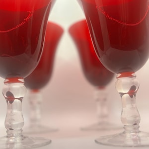 May include: A set of four wine glasses featuring deep red bowls that transition to clear glass stems. Each stem has a distinct bulbous, multi-tiered design leading down to a circular clear glass base. The red glass is translucent and smooth.