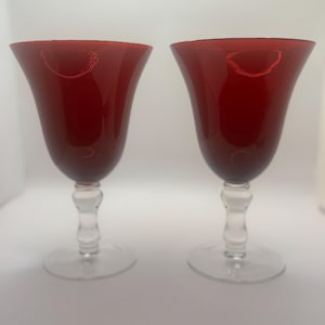 May include: A pair of matching wine glasses featuring deep red, flared bowls and clear, transparent stems with a rounded, decorative node detail. Each glass sits on a circular, clear glass base, showcasing a smooth, polished finish throughout.