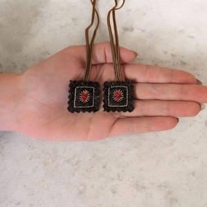 Pode incluir: Duas pequenas aplicações quadradas bordadas com um desenho de coração. As aplicações são cinza escuro com uma borda branca e um cordão castanho preso. O coração é vermelho com detalhes amarelos. As aplicações são seguradas numa mão.