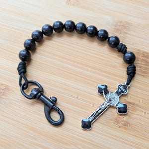 May include: A black beaded rosary with a silver crucifix and a black clasp. The crucifix has a detailed depiction of Jesus Christ. The beads are round and evenly spaced. The rosary is displayed on a wooden surface.