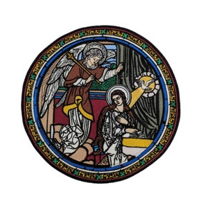 May include: A circular embroidered patch depicting a stained glass window scene. The scene shows an angel with wings, holding a staff, and a woman kneeling in prayer. The angel is wearing a brown robe with gold accents, and the woman is wearing a blue robe. The background is a dark blue and green stained glass window.