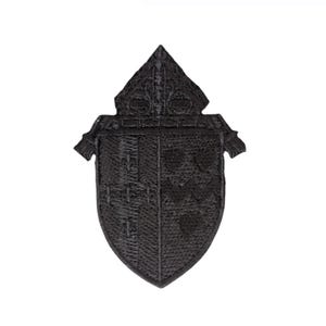 May include: A black embroidered patch in the shape of a heraldic shield, with a detailed design and a pointed top. The patch has decorative elements and is set against a white background.