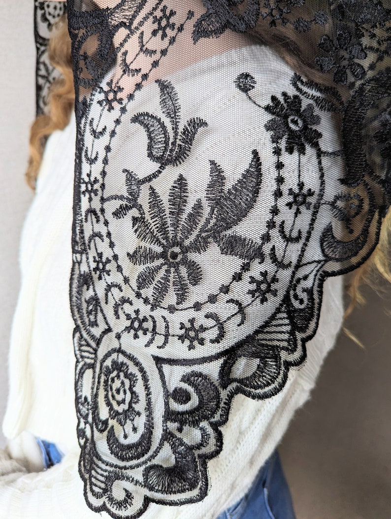May include: Black lace shawl with a floral pattern. The lace is delicate and intricate, with a scalloped edge.