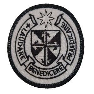 Puede incluir: Parche blanco bordado con un escudo blanco y negro, una estrella y el texto "Audare et Benedicere" y "The Academy" alrededor del escudo.