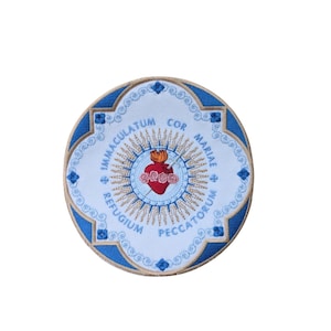 Puede incluir: Un parche circular blanco con un borde azul y una imagen bordada en oro del Corazón Inmaculado de María. El texto alrededor de la imagen dice "COR MARIAE IMMACULATUM REFUGIUM PECCATORUM".