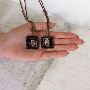 May include: Two small, square pendants with brown cords are displayed. Each pendant has a dark brown background with a decorative border. One pendant features gold-colored initials, while the other has a small image. The pendants are held in a hand.
