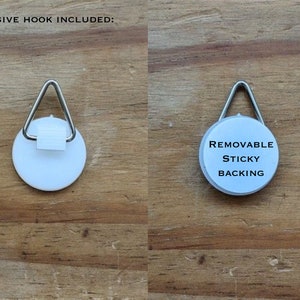 Może przedstawiać: Dwa obrazy białego haczyka samoprzylepnego z metalową pętlą tr&oacute;jkątną. Pierwszy obraz przedstawia haczyk z tekstem "ADHESIVE HOOK INCLUDED:". Drugi obraz przedstawia tył haczyka z tekstem "REMOVABLE STICKY BACKING".