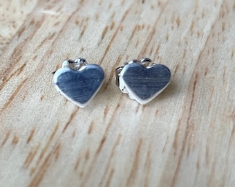 May include: A pair of small, silver heart-shaped stud earrings. The earrings are displayed on a light-colored wooden surface. The earrings have a simple, polished design, reflecting the light.