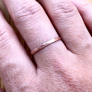 May include: A hammered copper ring is worn on a finger. The ring has a textured surface and a simple, minimalist design. The warm, reddish-brown color of the copper contrasts with the skin tone.