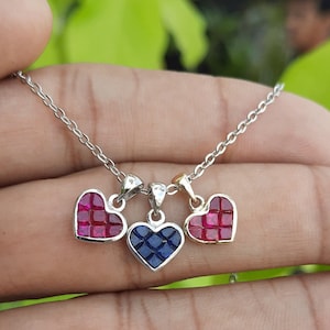 May include: A silver chain necklace with three heart-shaped pendants. The pendants are made of silver and have red and blue gemstones.