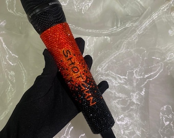 Könnte beinhalten: Ein schwarzes Mikrofon mit einem Farbverlauf aus orangefarbenen und schwarzen Strasssteinen. Der Text "SHOT ZN" ist auf dem Mikrofon zu sehen. Das Mikrofon wird von einer behandschuhten Hand gehalten.