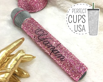 May include: Pink microphone covered in pink glitter with the word "Queen" spelled out in rhinestones. The microphone is on a white fluffy surface.