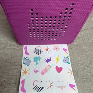 May include: A purple speaker with a white speaker grille and a sticker sheet with various colorful designs, including a crown, a heart, a star, a diamond, a shoe, a purse, a phone, a lightning bolt, a boombox, and a flower.