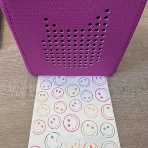 May include: A purple fabric box with a speaker grille in the shape of a heart. The box is sitting on a white sheet of paper with a pattern of colorful smiley faces.