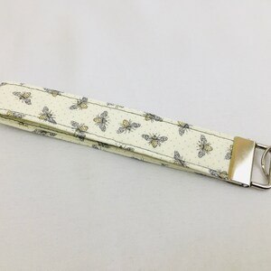 May include: A white fabric key chain with a silver key ring. The fabric has a pattern of small yellow and gray bees.